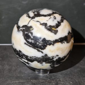 May include: A polished spherical stone featuring a high-contrast pattern of swirling black and white bands with subtle cream-colored inclusions. The smooth, rounded object rests on a small, clear circular stand, showcasing its natural mineral texture.