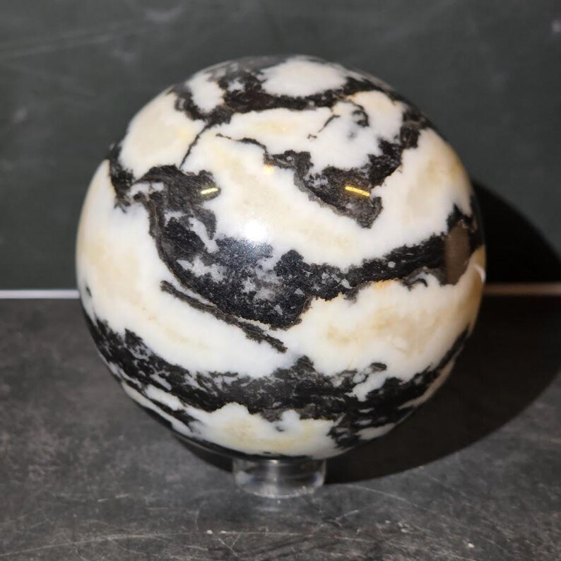 May include: A polished spherical stone featuring a high-contrast pattern of swirling black and white bands with subtle cream-colored inclusions. The smooth, rounded object rests on a small, clear circular stand, showcasing its natural mineral texture.