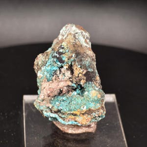 May include: A close-up of a mineral specimen with a rough, irregular shape. The rock features a mix of colors, including vibrant turquoise, brown, black, and hints of pink and yellow. The mineral appears to be crystalline in some areas.