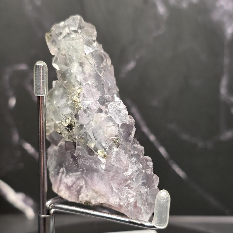 Puede incluir: Un grupo de cristales de fluorita natural con una mezcla de estructuras cristalinas de color p&uacute;rpura p&aacute;lido y transparente. La muestra mineral se exhibe sobre un soporte plateado, con un fondo oscuro y veteado.