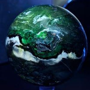Peut inclure: Une sph&egrave;re polie avec un motif tourbillonnant de verts profonds, de blancs et de noirs. Le globe en pierre pr&eacute;cieuse a une finition brillante, avec des zones de couleurs claires et sombres contrast&eacute;es. La sph&egrave;re est pr&eacute;sent&eacute;e sur un petit socle transparent.