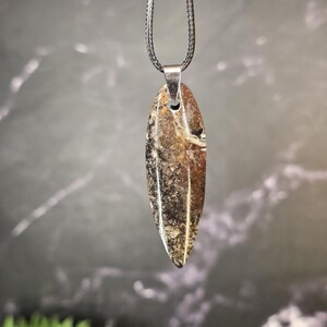 K&ouml;nnte beinhalten: Ein Anh&auml;nger mit einem langen, spitz zulaufenden, blattf&ouml;rmigen Stein mit gesprenkelter Textur in Braun-, Schwarz- und Cremet&ouml;nen. Der Stein h&auml;ngt an einer silberfarbenen Metall&ouml;se an einer d&uuml;nnen, geflochtenen dunkelgrauen Kordelkette.