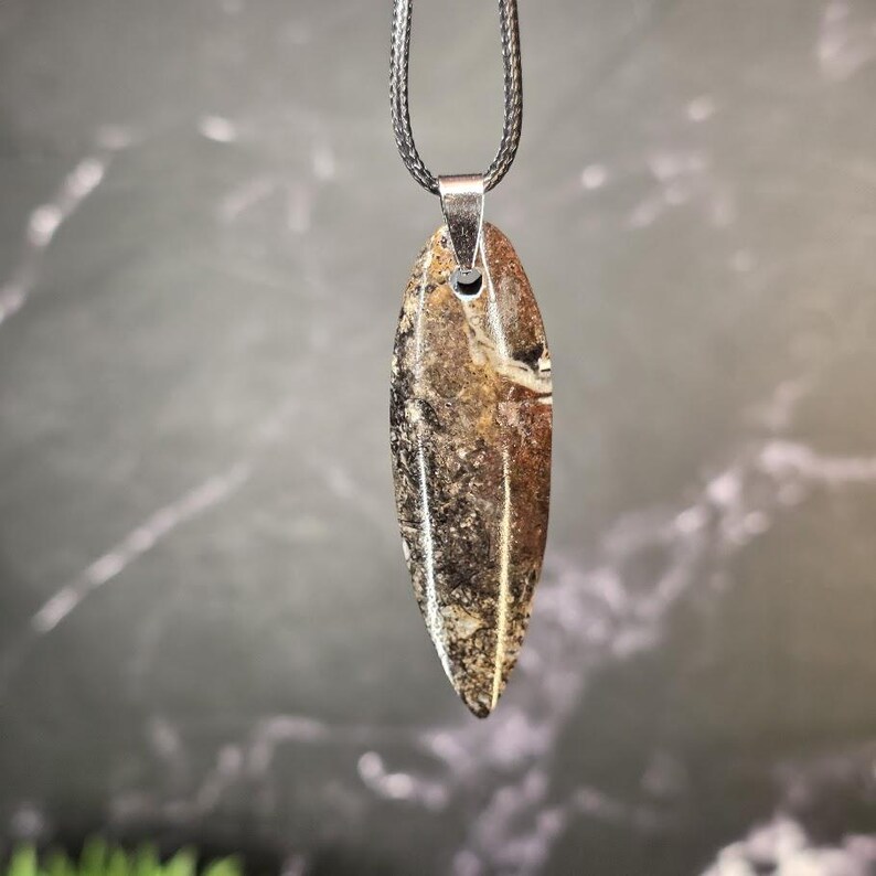 K&ouml;nnte beinhalten: Ein Anh&auml;nger mit einem langen, spitz zulaufenden, blattf&ouml;rmigen Stein mit gesprenkelter Textur in Braun-, Schwarz- und Cremet&ouml;nen. Der Stein h&auml;ngt an einer silberfarbenen Metall&ouml;se an einer d&uuml;nnen, geflochtenen dunkelgrauen Kordelkette.