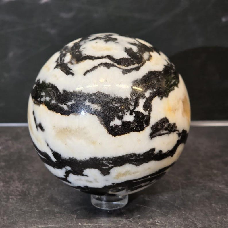 May include: A polished spherical stone featuring a high-contrast pattern of swirling black and white bands. The surface of the orb is smooth and reflective, showcasing organic, flowing lines that wrap around the entire rounded form of the object.