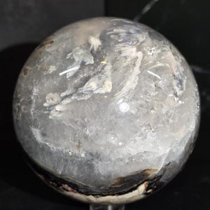 Peut inclure: Une sph&egrave;re de quartz clair avec des motifs internes blancs, gris et noirs. La boule de cristal polie a une surface lisse et repose sur un support transparent. La sph&egrave;re est un objet d&eacute;coratif.
