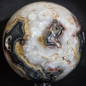May include: A polished agate sphere with a mix of colors, including white, black, brown, and gold. The sphere has a natural, organic pattern with visible layers and crystalline structures. The gemstone is displayed on a clear stand.