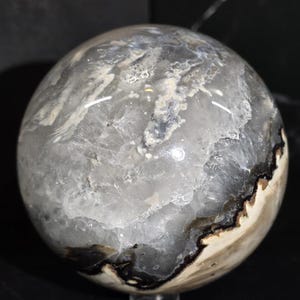 Peut inclure: Une sph&egrave;re de pierre polie avec un m&eacute;lange de motifs gris, blancs et noirs. L'orbite a une surface lisse et r&eacute;fl&eacute;chissante avec une apparence naturelle et organique. L'int&eacute;rieur de la pierre r&eacute;v&egrave;le des formations cristallines et une bordure sombre et contrast&eacute;e.