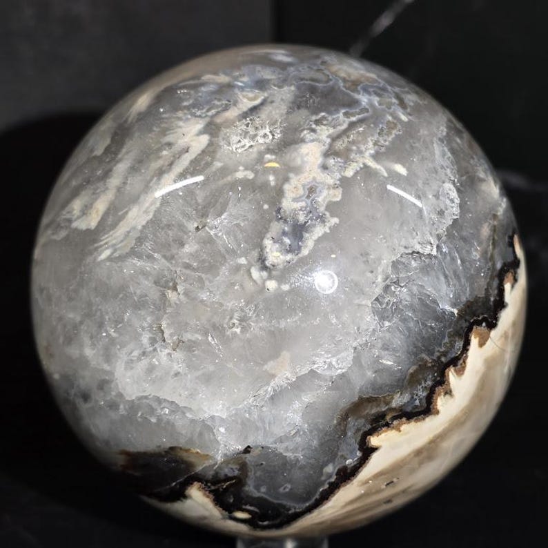 Peut inclure: Une sph&egrave;re de pierre polie avec un m&eacute;lange de motifs gris, blancs et noirs. L'orbite a une surface lisse et r&eacute;fl&eacute;chissante avec une apparence naturelle et organique. L'int&eacute;rieur de la pierre r&eacute;v&egrave;le des formations cristallines et une bordure sombre et contrast&eacute;e.