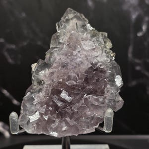 Puede incluir: Un grupo de cristales de fluorita natural con una mezcla de estructuras cristalinas moradas y claras. La muestra mineral est&aacute; montada sobre un soporte transparente contra un fondo oscuro, mostrando sus facetas geom&eacute;tricas y variaciones de color.