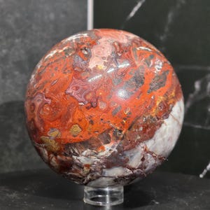 May include: A polished spherical stone featuring a complex pattern of deep red, burnt orange, white, and dark brown swirls. The surface displays intricate banding and mineral inclusions throughout the orb, which rests on a small clear circular stand.