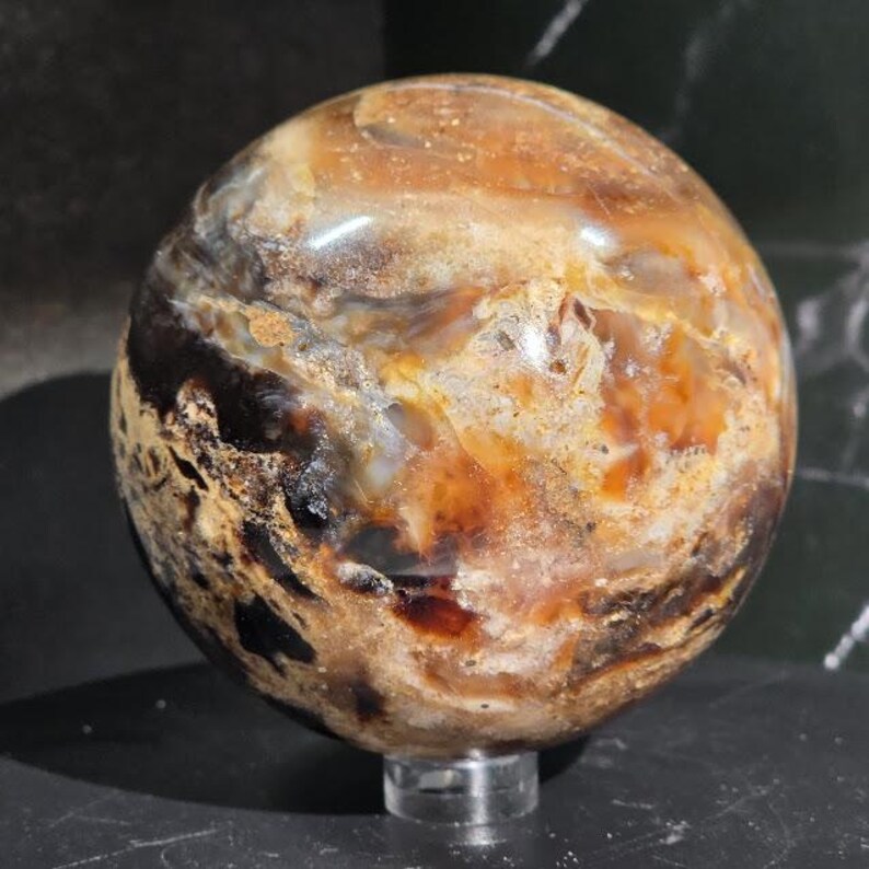 May include: A polished gemstone sphere with a swirling pattern of brown, tan, and amber hues. The orb is displayed on a clear acrylic stand, showcasing its natural colors and textures. This decorative sphere is a unique home decor item.