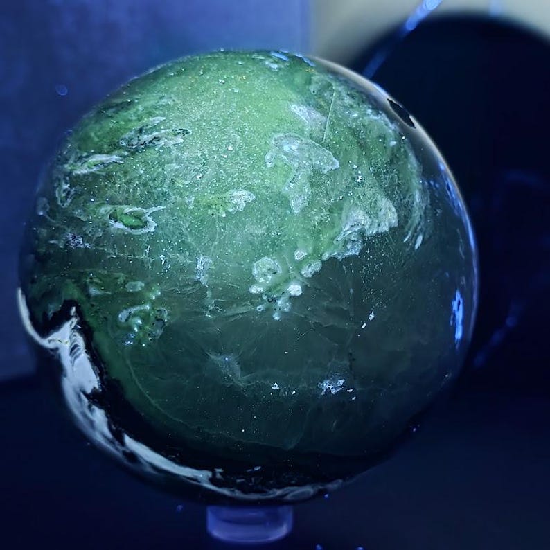 Peut inclure: Un objet sph&eacute;rique translucide avec un motif vert et blanc mouchet&eacute;. L'orbite semble &ecirc;tre faite d'une pierre ou d'un cristal poli, avec des veines et des inclusions internes visibles. La sph&egrave;re est expos&eacute;e sur une petite base bleue.