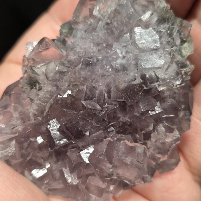 Puede incluir: Primer plano de un cristal de fluorita en bruto. El mineral exhibe una mezcla de tonos morados y claros, con una estructura geom&eacute;trica c&uacute;bica. La superficie refleja la luz, resaltando las facetas naturales. Aspecto cristalino y texturizado.