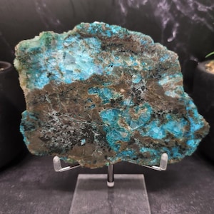 May include: A rough-cut, natural stone slab with a mix of teal, brown, and black colors. The stone is displayed on a clear stand, showcasing its unique texture and patterns. The overall aesthetic is earthy and organic.