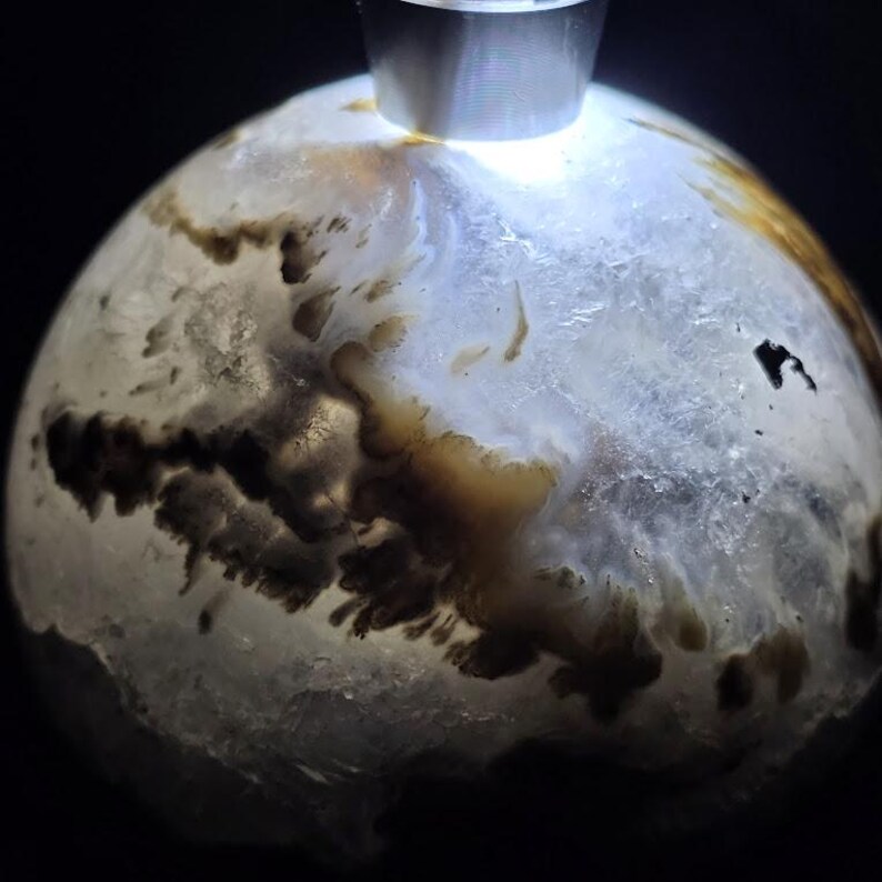 Peut inclure: Gros plan sur une pierre pr&eacute;cieuse sph&eacute;rique polie avec une base blanche translucide. La pierre pr&eacute;sente des motifs complexes d'inclusions brunes et noires, cr&eacute;ant un design naturel unique. Une pi&egrave;ce m&eacute;tallique argent&eacute;e est fix&eacute;e au sommet.