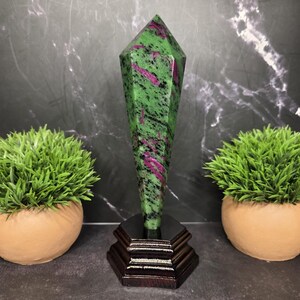 May include: A polished, faceted ruby zoisite crystal tower with vibrant green, black, and magenta-pink inclusions. The stone is mounted vertically on a dark brown, tiered wooden base, showcasing a sharp, pointed top and a smooth, reflective surface.