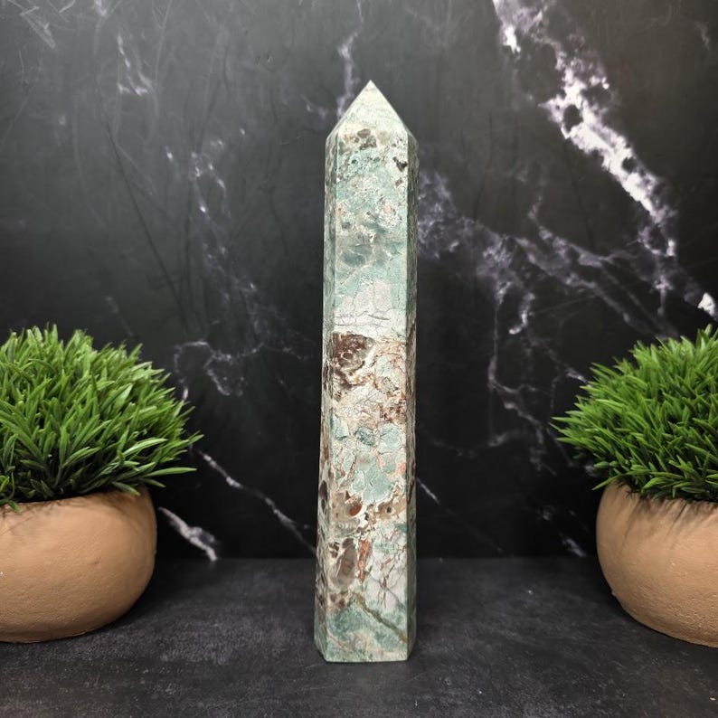 May include: A tall, six-sided crystal tower featuring a polished surface with a mottled pattern of sage green, cream, and dark brown mineral inclusions. The stone has a pointed, faceted top and a flat base, displaying natural variations in color.