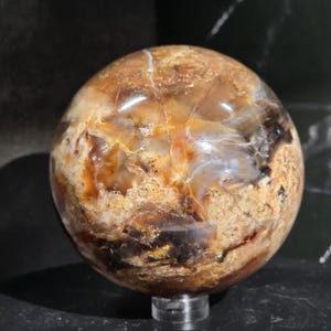 May include: A polished, spherical gemstone with a mix of brown, tan, and cream colors. The orb has a natural, marbled appearance with visible patterns and variations in color. It sits on a clear, circular stand, showcasing its unique texture and design.