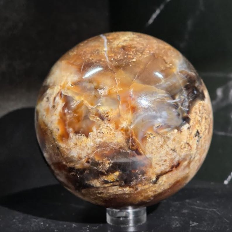 May include: A polished, spherical gemstone with a mix of brown, tan, and cream colors. The orb has a natural, marbled appearance with visible patterns and variations in color. It sits on a clear, circular stand, showcasing its unique texture and design.
