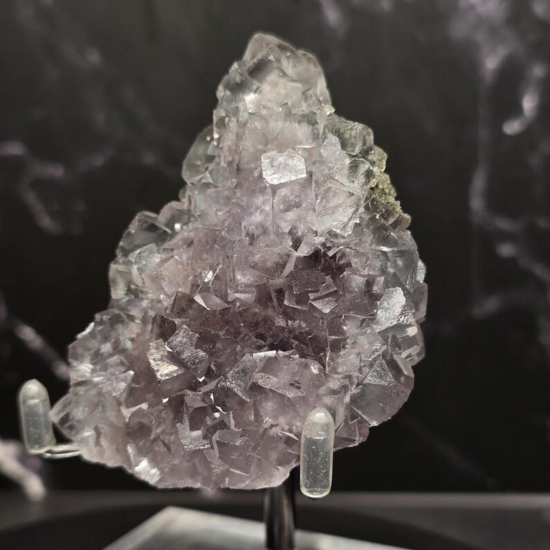 Puede incluir: Un grupo de cristales de fluorita morados y transparentes. Los cristales tienen una estructura c&uacute;bica y est&aacute;n montados en un expositor. La fluorita tiene un aspecto transl&uacute;cido con variaciones de color, que van del lavanda claro a tonos morados m&aacute;s profundos.