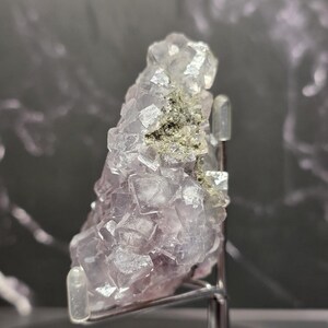 Puede incluir: Un grupo de cristales de fluorita natural con una mezcla de formaciones c&uacute;bicas de color p&uacute;rpura p&aacute;lido y transparente. El cristal tiene un peque&ntilde;o parche de inclusiones minerales de color verde gris&aacute;ceo. La muestra est&aacute; montada sobre un soporte met&aacute;lico.