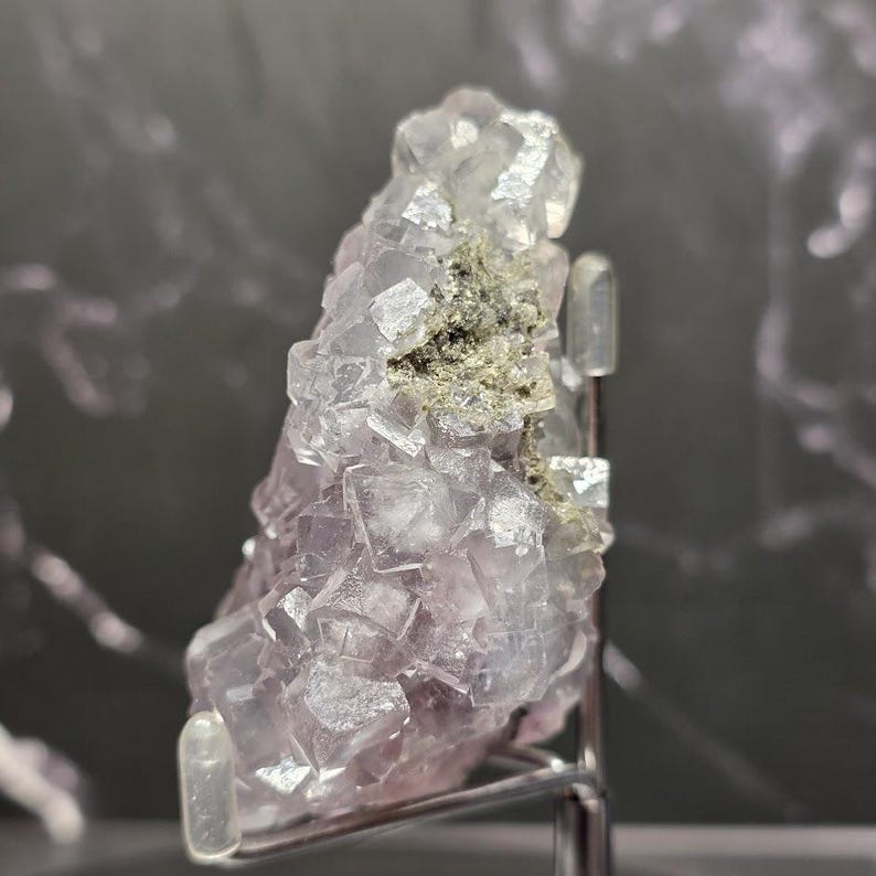 Puede incluir: Un grupo de cristales de fluorita natural con una mezcla de formaciones c&uacute;bicas de color p&uacute;rpura p&aacute;lido y transparente. El cristal tiene un peque&ntilde;o parche de inclusiones minerales de color verde gris&aacute;ceo. La muestra est&aacute; montada sobre un soporte met&aacute;lico.