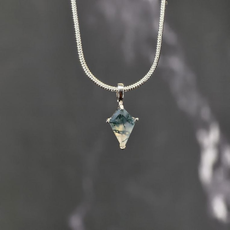 Puede incluir: Un collar de plata con un colgante en forma de diamante. El colgante presenta una piedra natural con patrones verdes y blancos. La cadena es de plata fina y texturizada. El colgante est&aacute; suspendido de una peque&ntilde;a fianza de plata.