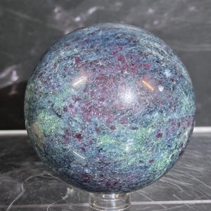 May include: A polished spherical mineral specimen featuring a mottled texture with deep blue, dark green, and vibrant magenta-red inclusions throughout the surface. The stone displays a crystalline structure with reflective highlights and sits on a clear base.