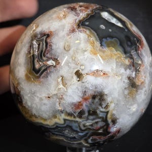 May include: A polished agate sphere with a complex pattern of white, black, brown, and orange bands. The gemstone ball has a smooth, rounded surface and is held by a hand. The sphere is a decorative item.