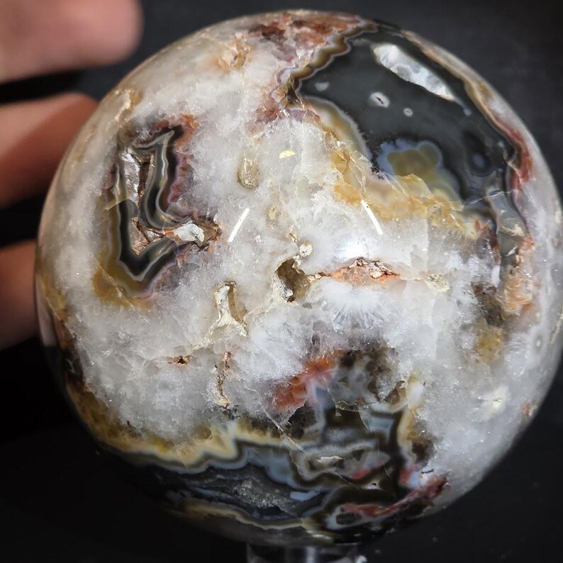 May include: A polished agate sphere with a complex pattern of white, black, brown, and orange bands. The gemstone ball has a smooth, rounded surface and is held by a hand. The sphere is a decorative item.