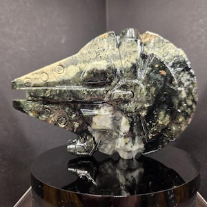 May include: A detailed sculpture of the Millennium Falcon starship, carved from a stone with green, black, and tan marbling. The model sits on a black pedestal, showcasing the ship's iconic design.