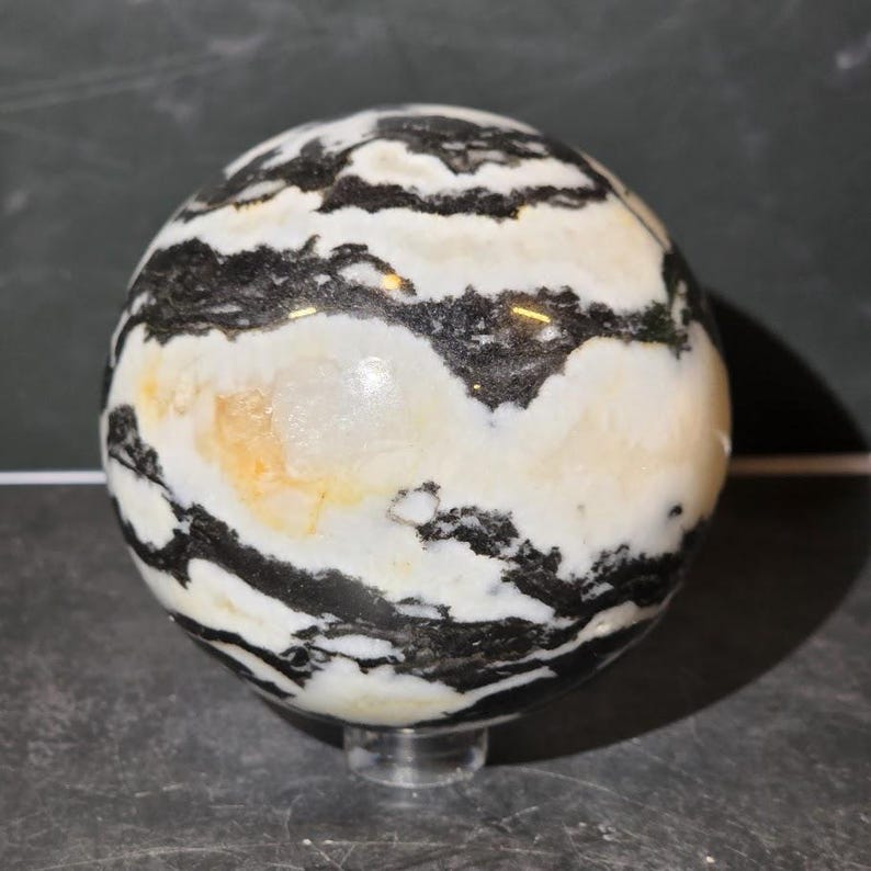 May include: A polished stone sphere featuring a high-contrast pattern of thick, irregular black bands alternating with creamy white and soft yellow mineral layers. The surface is smooth and reflective, showcasing natural geological banding throughout.