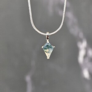 Puede incluir: Un collar de plata con un colgante en forma de diamante. El colgante presenta una piedra verde y blanca, y la cadena tiene un dise&ntilde;o texturizado y tejido. El fondo es gris liso.