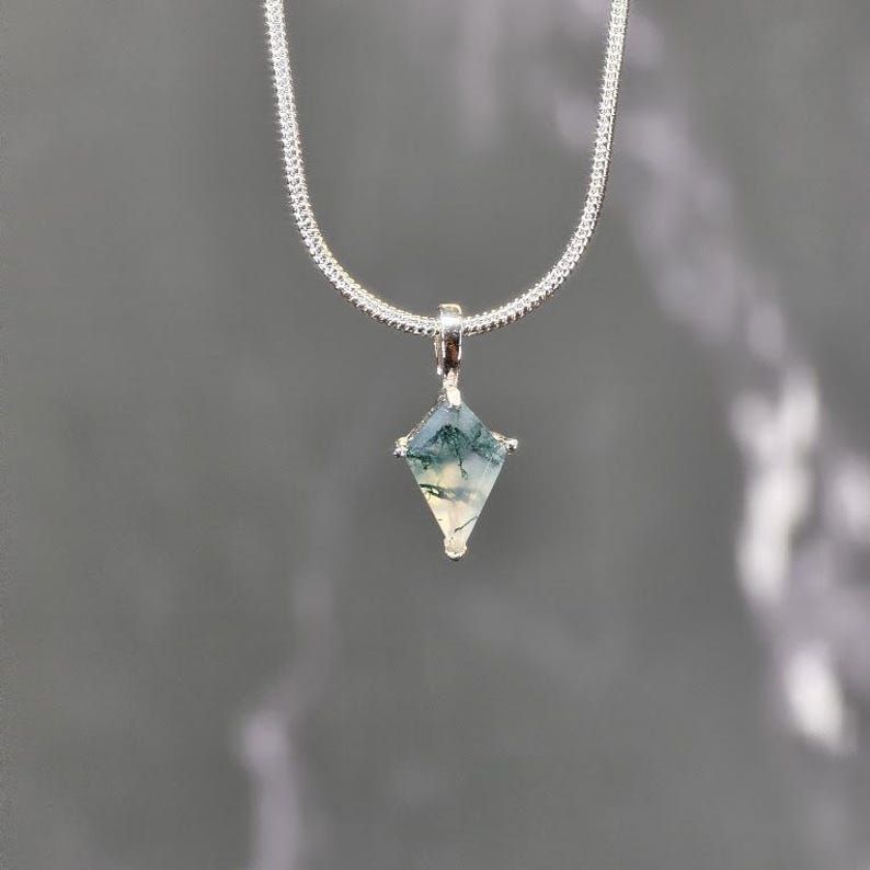 Puede incluir: Un collar de plata con un colgante en forma de diamante. El colgante presenta una piedra verde y blanca, y la cadena tiene un dise&ntilde;o texturizado y tejido. El fondo es gris liso.