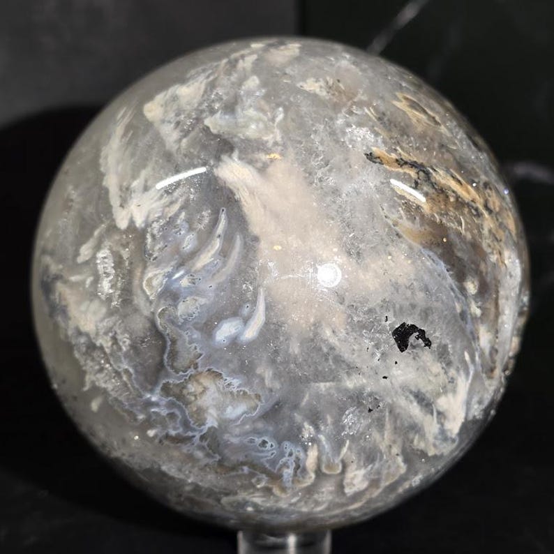 Peut inclure: Une boule de cristal sph&eacute;rique et transparente avec des motifs internes blancs, gris et bruns. La surface polie refl&egrave;te la lumi&egrave;re, mettant en valeur les formations naturelles complexes &agrave; l'int&eacute;rieur de la pierre. La sph&egrave;re est mont&eacute;e sur un support transparent.