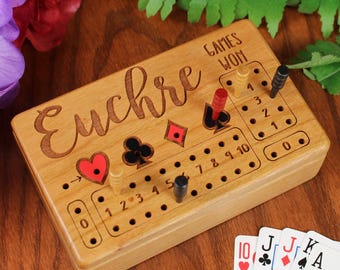 May include: A rectangular wooden Euchre scorekeeper board with engraved card suit symbols, numbered holes, and four colored pegs. The board features the text "Euchre" and "Games Won" with a set of playing cards showing a ten, jack, jack, and king.