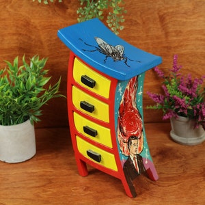 May include: A small, curved wooden chest with five drawers painted in bright colors. The top drawer is blue with a painted image of a fly. The other drawers are yellow with black handles. The side of the chest is painted with a red and yellow flame design and a man's face.