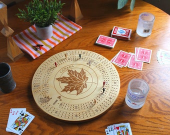 Könnte beinhalten: Ein hölzernes Cribbage-Spielbrett mit Ahornblatt-Design und dem Wort "CANADA", umgeben von Spielkarten, einem Kartenspiel und Schnapsgläsern. Eine kleine Topfpflanze und ein Kerzenhalter sind ebenfalls vorhanden.