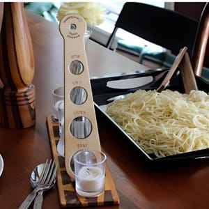 May include: Wooden pasta measuring tool with four different lengths of pasta strands. The tool is labelled "one", "two", "three", and "four". There are bowls of cooked pasta on a wooden table.