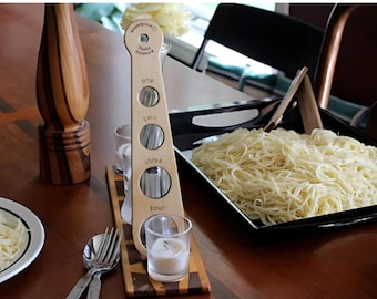 May include: Wooden pasta measuring tool with four different lengths of pasta strands. The tool is labeled "one", "two", "three", and "four". There are bowls of cooked pasta on a wooden table.