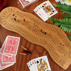 May include: A wooden cribbage board with a wavy shape, featuring numbered tracks, a start and finish line, and small coloured pegs. The board is surrounded by several playing cards with red and black suits and a deck box on a wooden surface.