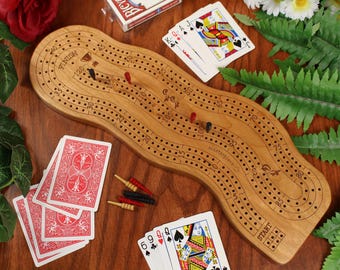May include: A wooden cribbage board with a wavy shape, featuring numbered tracks, a start and finish line, and small colored pegs. The board is surrounded by several playing cards with red and black suits and a deck box on a wooden surface.