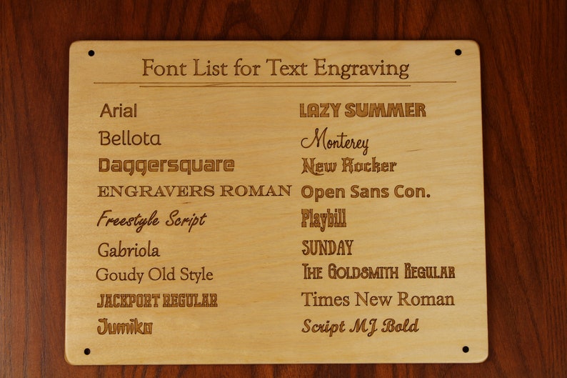 May include: A rectangular light-colored wooden plaque featuring the engraved title Font List for Text Engraving. The board displays two columns of various font names, including Arial, Bellota, Daggersquare, Engravers Roman, Freestyle Script, Gabriola, Goudy Old Style, Jackport Regular, Jumiko, Lazy Summer, Monterey, New Rocker, Open Sans Con., Playbill, Sunday, The Goldsmith Regular, Times New Roman, and Script MJ Bold.