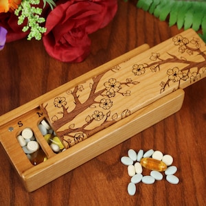 May include: A wooden pill box with a sliding lid, featuring a carved cherry blossom branch design. The box is open, revealing various pills. A small pile of pills sits next to the box. The box is on a wooden surface, with flowers in the background.