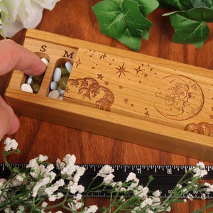 May include: Wooden pill box with a moon and stars design. The box has seven compartments labelled S, M, T, W, T, F, and S.