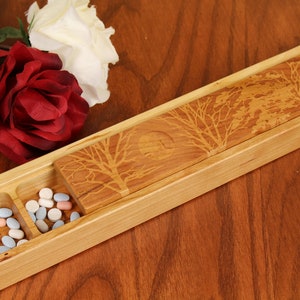 May include: A wooden box with a sliding lid, featuring a laser-etched design of trees and a moon. The box is divided into two compartments, each containing a variety of colourful pills.
