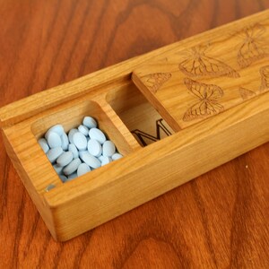 May include: A wooden pill box with a hinged lid. The lid is decorated with a pattern of butterflies. The box has two compartments, one of which is filled with light blue pills.