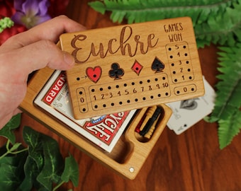 May include: A rectangular wooden Euchre scorekeeping board with a sliding lid. The board features engraved card suit symbols, a numbered scoring track from zero to ten, and a section labeled Games Won. A deck of playing cards and two pegs are inside.
