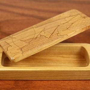 Peut inclure: Une petite boîte en bois avec un couvercle à charnière. Le couvercle est ouvert et présente un motif de feuilles sculptées. La boîte est en bois clair et a une finition naturelle.