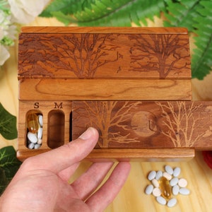 May include: Wooden pill box with a sliding drawer and two compartments labelled "S" and "M". The box is decorated with an engraved design of trees and a moon.